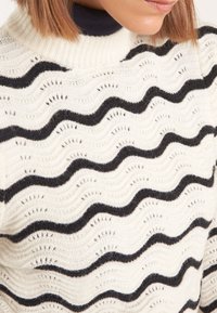 Pull en maille crème avec un motif ondulé texturé en noir, doté d'un col montant et de détails complexes sur tout le tissu.