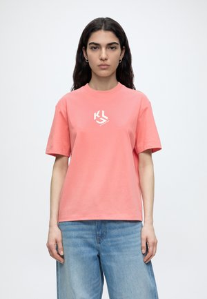Femme aux longs cheveux foncés portant un t-shirt corail à manches courtes avec un logo blanc et un jean bleu clair, debout devant un fond clair uni.