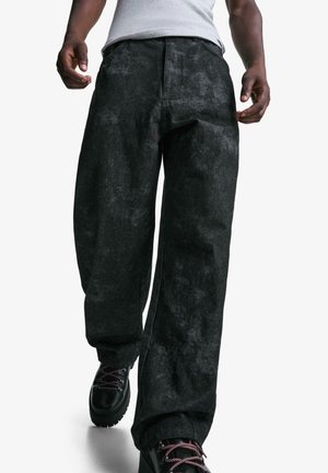 Osoba nosząca luźne czarne spodnie i czarne buty sznurowane z białym bezrękawnikiem, idzie do przodu na białym tle.