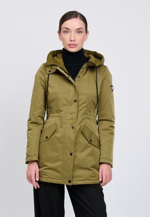 Frau mit heller Haut und dunklen Haaren trägt eine olivgrüne Kapuzenjacke und schwarze Hose vor einem einfarbigen hellen Hintergrund.