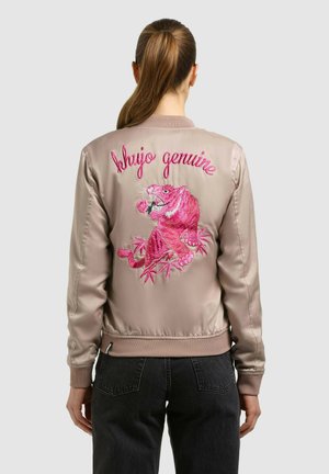 Nő bézs színű szatén bomber dzsekiben, amelynek hátán rózsaszín hímzett tigris látható, és egy rózsát tart, valamint a "shujo genuine" felirat olvasható rajta.