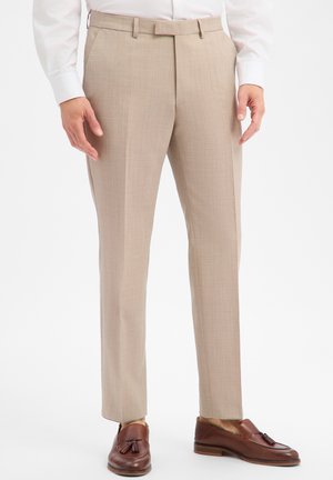 Man draagt beige op maat gemaakte broek, bruine leren loafers met kwastjes en een wit overhemd met lange mouwen tegen een witte achtergrond.