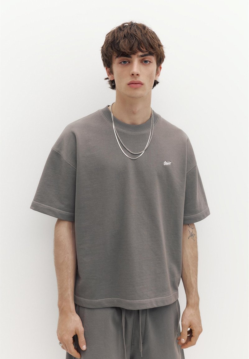 Jeune homme aux cheveux courts bruns portant un t-shirt gris ample et un pantalon assorti, des chaînes argentées, et un petit tatouage sur l'avant-bras gauche.