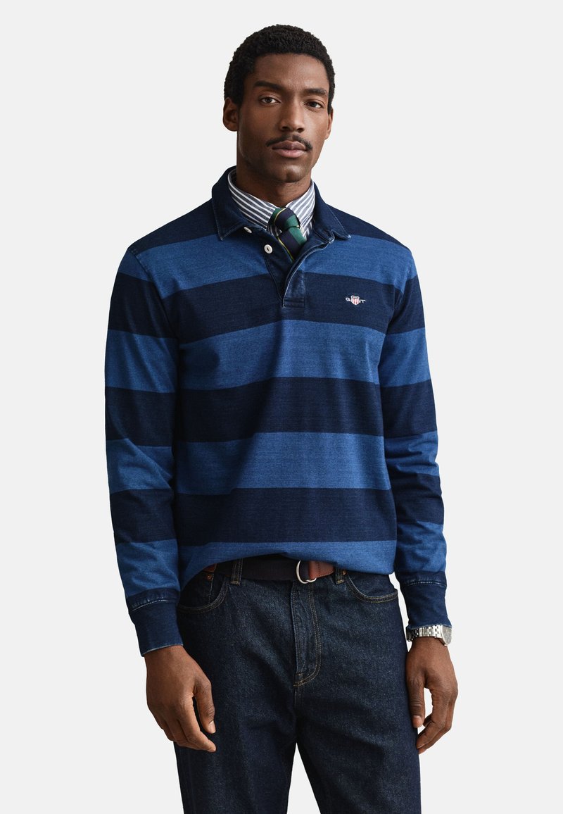 Uomo che indossa una maglia da rugby a maniche lunghe a righe blu e blu navy sopra una camicia a righe con colletto, cravatta verde e blu, jeans scuri e orologio d'argento.