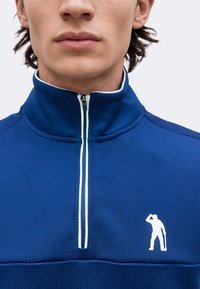 Jeune homme portant un haut de sport bleu à fermeture éclair avec un petit logo blanc en silhouette d'une personne tenant un bâton sur la poitrine.