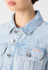 Lyseblå denimjakke med sølvfargede trykknapper, klassisk krage og en frontlomme med en etikett som sier "Calvin Klein."