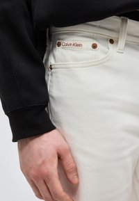 Αριστερό χέρι ακουμπισμένο δίπλα σε λευκό τζιν με ετικέτα Calvin Klein στην μικρή μπροστινή τσέπη, σε συνδυασμό με μαύρο ρούχο με μακριά μανίκια.