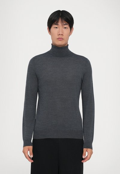Ung mand iført en mørkegrå tætsiddende rullekravesweater og sorte bukser, stående mod en ensfarvet hvid baggrund.