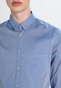 Modrá pruhovaná košile na knoflíky s špičatým límcem, náprsní kapsou a hladkou strukturou. Knoflíky jsou světlé a kulaté.
