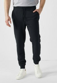 Pantalones de chándal negros con cintura ajustable, bolsillos laterales y tobillos con puños. Llevado con zapatillas blancas sobre un fondo neutral.