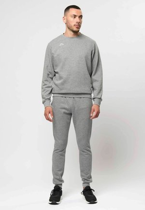 Čovjek stoji, nosi sivu majicu s dugim rukavima i odgovarajuće sivke trenirke s crnim tenisicama, gleda ulijevo na jednostavnoj pozadini.