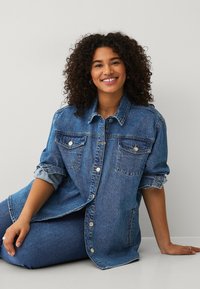 Giacca di jeans blu oversize con due tasche sul petto, chiusura con bottoni e maniche arrotolate, abbinata a jeans blu coordinati.