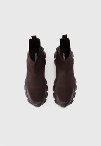 Tmavě hnědé slip-on tenisky s pleteným svrškem, texturovanými částmi a silnou, robustní podrážkou. Mají poutko pro snadné obouvání.