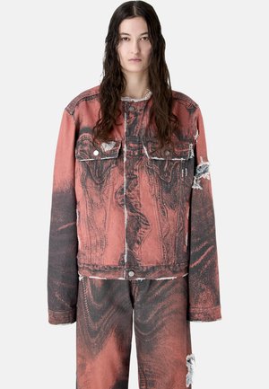 Kobieta z długimi ciemnymi włosami, ubrana w przetartą, dużą czerwoną i czarną kurtkę jeansową oraz pasujące spodnie, stojąca na jednolitym tle.