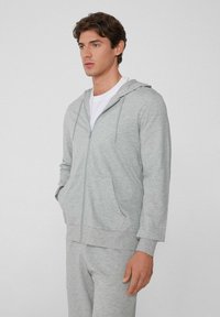Mann in heller grauer Zip-Up-Hoodie und passender Jogginghose, der vor einem schlichten Hintergrund steht, mit den Händen in den Taschen.