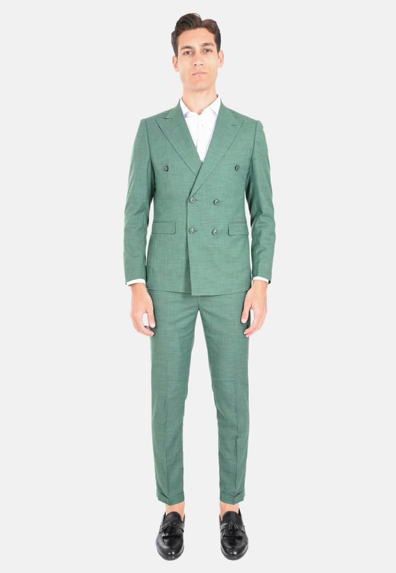 Completo verde doppiopetto in tessuto testurizzato, slim fit, con pantaloni abbinati. Presenta scarpe scure e una camicia bianca sotto.