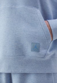 Hellblaues Hoodie aus weichem Stoff, mit einer Kängurutasche und einem strukturierten Patch mit einem grafischen Logo in der unteren Ecke.