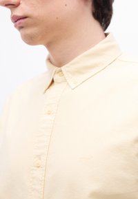 Jeune personne portant une chemise jaune pâle à boutons avec un col, partiellement visible du cou jusqu'au milieu de la poitrine.