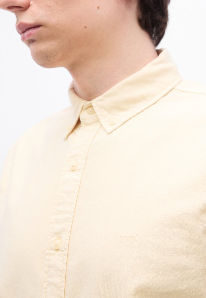 Jeune personne portant une chemise jaune pâle à boutons avec un col, partiellement visible du cou jusqu'au milieu de la poitrine.