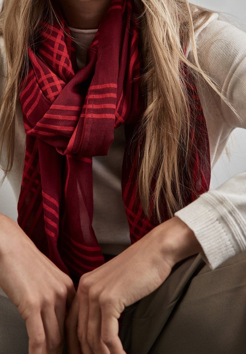 Personne portant une chemise blanche à manches longues et une écharpe rouge à motifs, avec de longs cheveux blonds et les mains reposant sur les genoux.