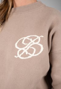 Pull en maille beige avec un motif de lettres blanches entrelacées sur la poitrine, porté par une personne aux longs cheveux châtain clair.