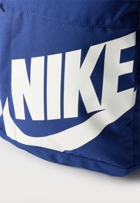 Μπλε υφασμάτινη τσάντα με μεγάλο λευκό λογότυπο NIKE και το σύμβολο ‘swoosh’ τυπωμένα στην εμπρόσθια τσέπη, εμφανιζόμενη σε κοντινή λεπτομέρεια.