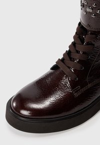 Burgundy patent leather ankle boot with a rounded toe, black rubber sole, and brown laces; features metal stud accents on ankle.