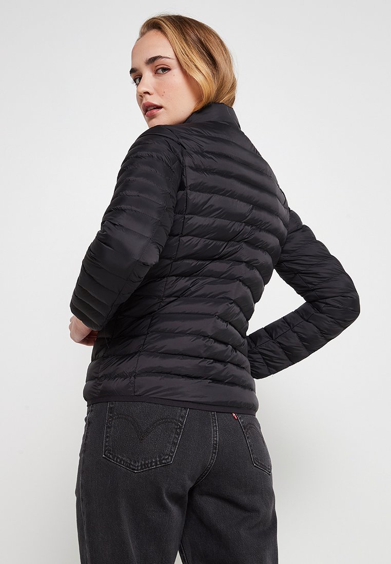 Doudoune noire avec un design ajusté, texture matelassée et col haut. Modèle portant un jean foncé, mettant en valeur la silhouette élégante de la doudoune.