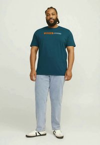 Homem com tranças a usar uma t-shirt Jack & Jones de teal escuro, jeans azul claro e ténis brancos com riscas pretas, de pé contra um fundo simples.