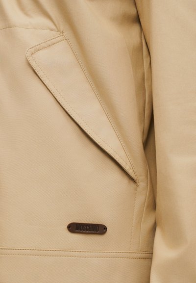 Τσέπη από μπεζ παλτό με λοξή τάπα και καφέ ορθογώνια ετικέτα με ανάγλυφο λογότυπο "MOSCHINO" ραμμένη κάτω από την τάπα της τσέπης.