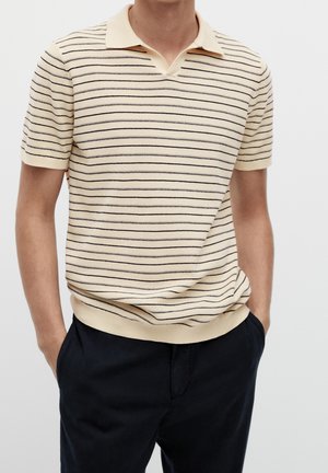 Mand iført en kortærmet beige polo med tynde sorte vandrette striber og sorte bukser, hænder i lommerne mod en ensfarvet baggrund.