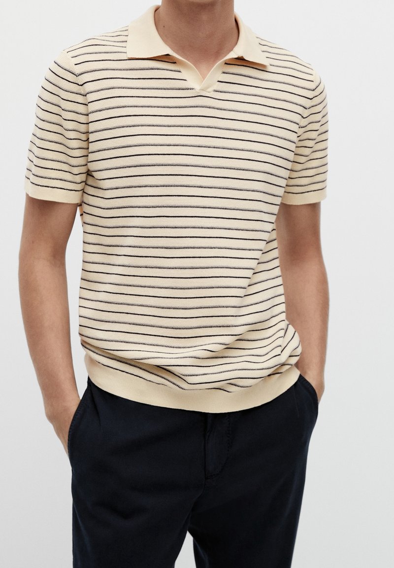 Homme portant un polo beige à manches courtes avec de fines rayures horizontales noires et un pantalon noir, les mains dans les poches devant un fond uni.