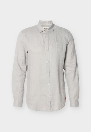 Gaiši pelēka garām piedurknēm pogājama krekls ar apkakli, vienu aproces pogu un nelielu sarkanu logotipu pie apakšējās malas uz baltas virsmas.