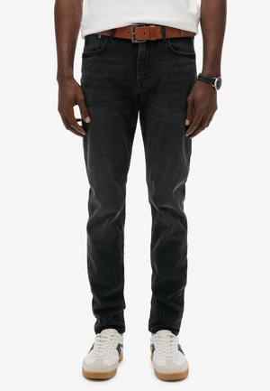 Mand iført sorte jeans, brun bælte, hvide sneakers, hvid skjorte og sort armbåndsur, står foran en hvid baggrund.