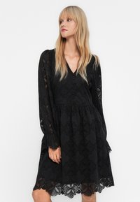 Blonde Frau, die ein schwarzes, kniebartiges Spitzenkleid mit langen Ärmeln, Wellenkante und V-Ausschnitt trägt, und zur Seite schaut vor einem weißen Hintergrund.