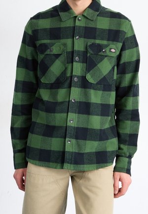 Chemise en flanelle à carreaux verts et noirs, avec un col, deux poches poitrine et une fermeture à boutons sur le devant. Texture douce et coupe standard.