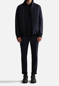 Homme portant une veste zippée bleu marine foncé, un pantalon assorti et des baskets noires, debout devant un fond blanc.