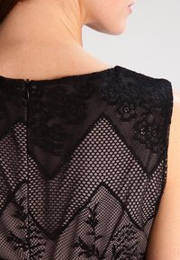 Žena s odhalenou horní částí zad a ramenem, oblečená v černém krajkovém oděvu s květinovými a geometrickými síťovými vzory, zapnutém malým černým zipem.