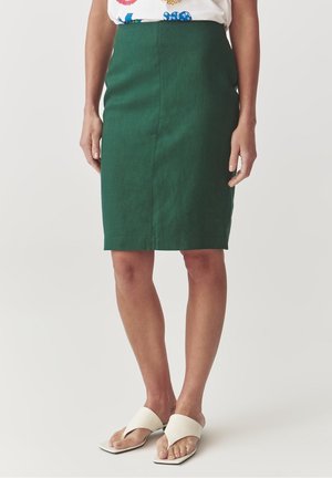 Mujer con falda lápiz verde oscuro hasta la rodilla, sandalias de tacón blancas abiertas en los dedos y blusa blanca con patrones florales coloridos.