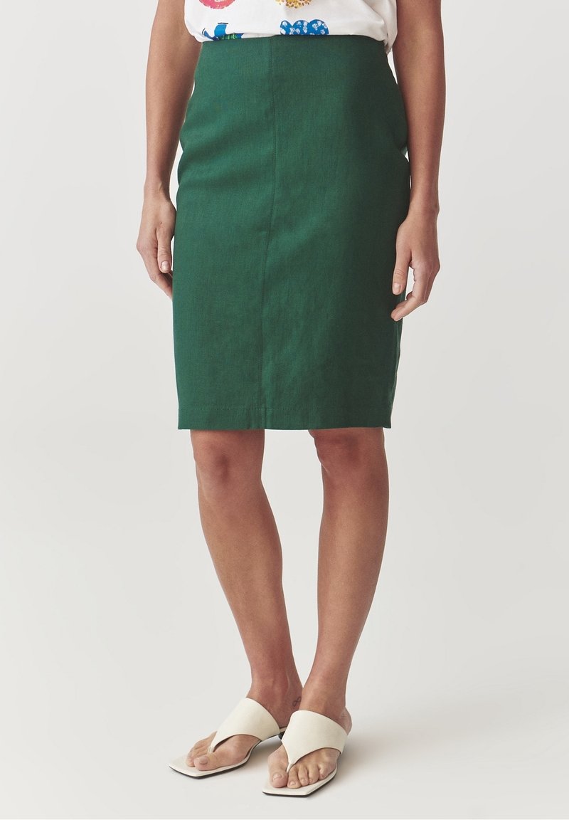 Femme portant une jupe crayon vert foncé jusqu'aux genoux, des sandales à talons blanches à bout ouvert, et un chemisier blanc à motifs floraux colorés.