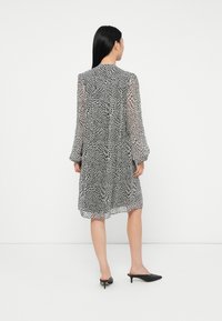 Γυναίκα με μακριά μαύρα μαλλιά, φορώντας ένα μαύρο και λευκό φόρεμα έως το γόνατο με σχέδιο και μαύρες γόβες με τακούνι, στέκεται απέναντι σε έναν/plain λευκό τοίχο.