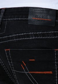 Poche arrière d'un jean en denim noir avec des surpiqûres grises et orange, et une étiquette en cuir noir de la marque Camp David au-dessus de la poche.
