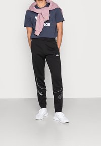 Černé tepláky s bílým lemováním a logem, v kombinaci s námořnickě modrým tričkem s velkým bílým logem a světle růžovou mikinou přehozenou přes krk.