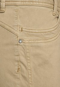 Beige cotton pants with a smooth texture, featuring contrasting stitching, a decorative silver button, and a curved pocket design.