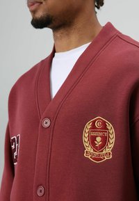 Bordeauxrode cardigan van zachte stof, met een v-halsontwerp, twee knopen en een geborduurd logo met een bloemmotief in goud.