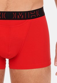 Boxer da uomo rossi realizzati in tessuto elasticizzato, con una cintura elastica nera che presenta un testo rosso. Design aderente con bordi senza cuciture e una superficie liscia.