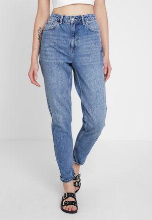 Femme portant un jean taille haute bleu clair avec les ourlets roulés, des sandales noires à doubles boucles et un crop top blanc, debout devant un fond uni.