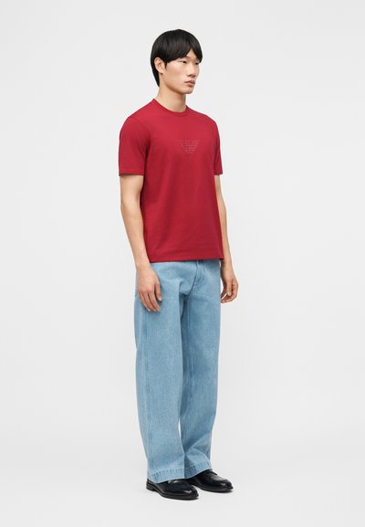 Άνδρας που στέκεται, φορώντας κόκκινο κοντομάνικο πουκάμισο με λογότυπο, ανοιχτό μπλε τζιν με φαρδιά πόδια και μαύρα παπούτσια, μπροστά σε απλό φωτεινό φόντο.