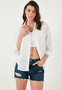White button-up shirt made of lightweight fabric, worn over a fitted top. Paired with distressed denim shorts, showcasing a casual look.