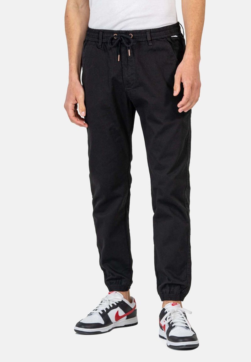Pantalones de chándal negros con cintura elástica y cordón. Presentan piernas ajustadas y dobladillos acabados, combinados con zapatillas negras y rojas.